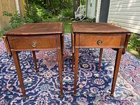 Pair of wooden side tables outdoors on patterned rug showing front view with drawer and drop leaf sides