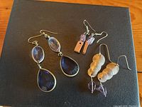 Three pairs of earrings displayed on a black surface showing the distinct styles and purple stones.