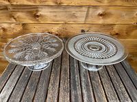 Two vintage glass cake plates displayed side by side on a wooden surface with a wood panel background, showing intricate pressed glass designs.