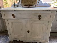 Front view of small white hutch showing two cabinet doors and three knobs on drawer with distressed, cracked paint finish.