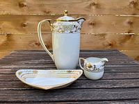 Full view of the three-piece tea set on a wooden table outside, showing the teapot, creamer, and tray with gold floral designs and white porcelain body.