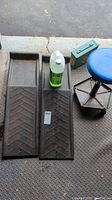 Two black plastic wheel ramps, partial bottle of green windshield washer fluid, metal first aid box, and blue seat adjustable shop stool on garage metal floor.