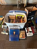 View of the full tote with books inside showing a collection of biographies, novels, and music books. Tote is light gray with blue lid, books in various sizes and colors.