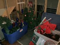 Photo showing blue storage bin with wreaths and other decorations along with several Christmas trees standing upright nearby.