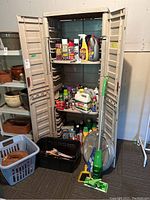 Full view of beige plastic storage cabinet with three shelves filled with assorted bottles and cans of cleaning and household products plus green Swiffer mop standing next to it. Laundry baskets and wooden basket placed in front on floor.