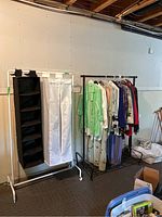 Two clothing racks side by side in a basement setting. Black metal rack holding hanging clothes; white tubing rack holds two hanging fabric storage pieces (one black open shelving organizer, one white zip-front organizer).