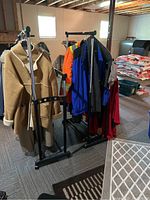 Two adjustable height two-bar clothing racks holding jackets and coats in a basement setting with visible ceiling beams and other stored items.