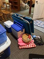 Wide shot showing folding cot, two coolers (Rubbermaid and Coleman), tablecloths, woven placemats, darts, playing cards, and black paper plate holders arranged on carpeted floor