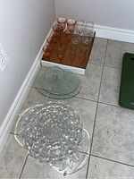 Overview of all glassware and trays included in lot placed on tile floor near wall baseboard.