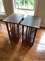 Pair of matching wooden end tables with two lower shelves, shown positioned by windows on wood floor. Visible wood grain and dark finish with some wear.