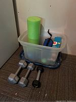 Photo showing green foam yoga roll, blue ankle weights, small weighted ball, and clear plastic storage bin with lid.