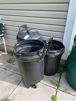 Three black Rubbermaid garbage pails; two have wheels and handles, one has a lid.