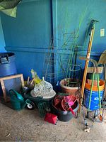 Photo shows garden stakes, tomato cages, plastic and metal planters, watering can, garden tools, and buckets grouped in a corner against a blue wall.