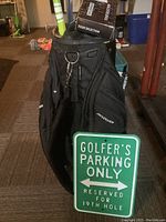Revolver black golf bag standing upright, showing multiple zippered pockets and top zipper with Bag Boy logo, with a golfer parking sign propped in front.