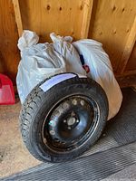 Four tires stacked with one visible showing mounted on a steel rim, inside storage with white bags behind.