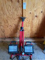 Two NOMA portable industrial lights placed on the ground with red adjustable tripod stand behind them, against plywood wall.