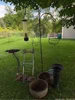 Overview photo showing two hanging bird feeders on a tall black metal stake with a sunburst trellis behind, wire basket feeder, metal folding chair, bird bath, cast iron lantern, metal bird decor on the ground, two large rusty metal bowls and other garden items.