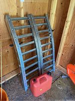 Pair of two metal car ramps leaning against a wooden wall inside a shed, showing length and basic structure with a red gas container in front.