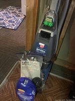 Front and side view of Hoover SteamVac Deluxe upright carpet cleaner with attached powered hand tool, bottle of filter cleansing solution, and user manual bag.
