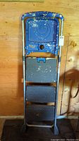 Full front view of blue 5-foot step ladder with three black anti-slip steps and paint splattered all over, resting against a wood wall.