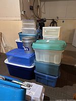 Stacked assortment of plastic storage bins of varying sizes and colors including blue, teal, clear, and white; includes a grey Lowe's pail with lid and handle on top of bins.