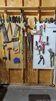 Wide view of pegboards holding multiple hand tools including saws, hammers, cutters, pliers, ropes, and cords in a garage setting.