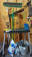 Photo showing yard care tools including large brooms, leaf and garden rakes, Friskers weed puller, ice melt bags, ice melt spreader, metal bucket, hand trimmers, scoops, trowels, trimmer line, and a plastic rack holding tools vertically.