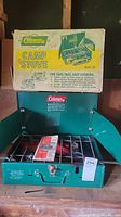 Photo showing Coleman camp stove Model 421 with original box behind it, metal green stove has two burners and fold-up side windshields.