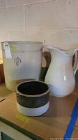 Stoneware crocks and ceramic pitcher side by side on a shelf, showcasing sizes and finishes.