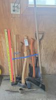 Full set of gardening tools leaning against wooden wall including axes, sledgehammer, mattock, shovel and two measuring rulers