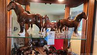 Five Beswick horse figurines displayed on glass shelves in a wooden cabinet. Central smaller horse has a broken leg.