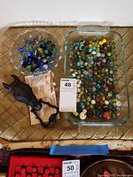 Decorative footed glass bowl with assorted marbles, and cast metal horse head door knocker on the left side on a cloth surface.