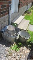 Outdoor photo of three nested rectangular galvanized metal bins and three galvanized metal pails placed beside a porch step on gravel and grass.