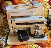 Front view of Kenmore sewing machine with foot pedal and carrying case on floral couch and crocheted blanket background.