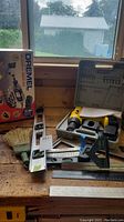 Photo of Dremel rotary tool kit box, cordless drill in case, levels, rulers, brush, and other measuring tools on a wooden workbench near a window.