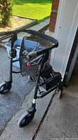 Front angle showing black metal frame walker with mesh basket and padded seat, as well as hand brakes and wheels.