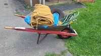 Side angle of loaded wheelbarrow showing tarps, garden hose, extra wheel inside, and red metal handles with wooden grips.