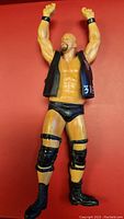 Full view of Stone Cold Steve Austin action figure with arms raised, wearing black vest and wrestling attire, displayed against a red background.