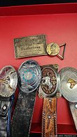 Four leather belts with decorative buckles showing wear including Budweiser and buffalo nickel designs.