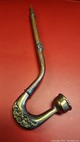 Full view of the curved brass pipe showing the bowl and engraved designs on the lower curved part.