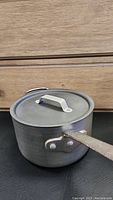 Aluminum cooking pot with lid and metal handle resting on a black surface in front of a wooden background.