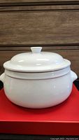 Front view of white ceramic Dutch oven with lid on red surface, showing round body and lid with knob.