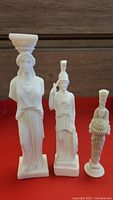 Front view of three alabaster statues on red surface with wooden background, showing Caryatid, Athena Pallas, and Artemis Diana of Ephesus from left to right.