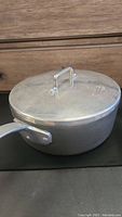 Side view of aluminum saucepan with lid and long handle on black surface in front of wooden furniture.
