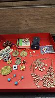 Full view of all assorted pins, keychains, necklace, earrings, and other small jewelry items arranged on red tray.