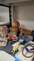 Photo showing various decorative and collectible items including carved wooden sphere, woven basket, crystal glassware, figurines, and beaded necklace.