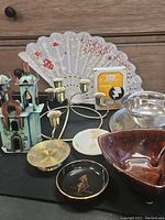 Photo of various miscellaneous decorative items including metal church figurine, multiple candle holders, ceramic bowls, and a hand fan on a black surface