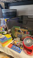 Mesh organizer with colored pencils, pens, push pins, and small red fan on table.
