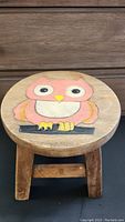 Top view of the round wooden stool seat showing painted pink owl with black eyes and yellow beak perched on black branch.