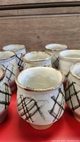 Eight vintage ceramic cups arranged on a red surface, showing the white glaze and brown crosshatch design with some visible wear and glaze irregularities.